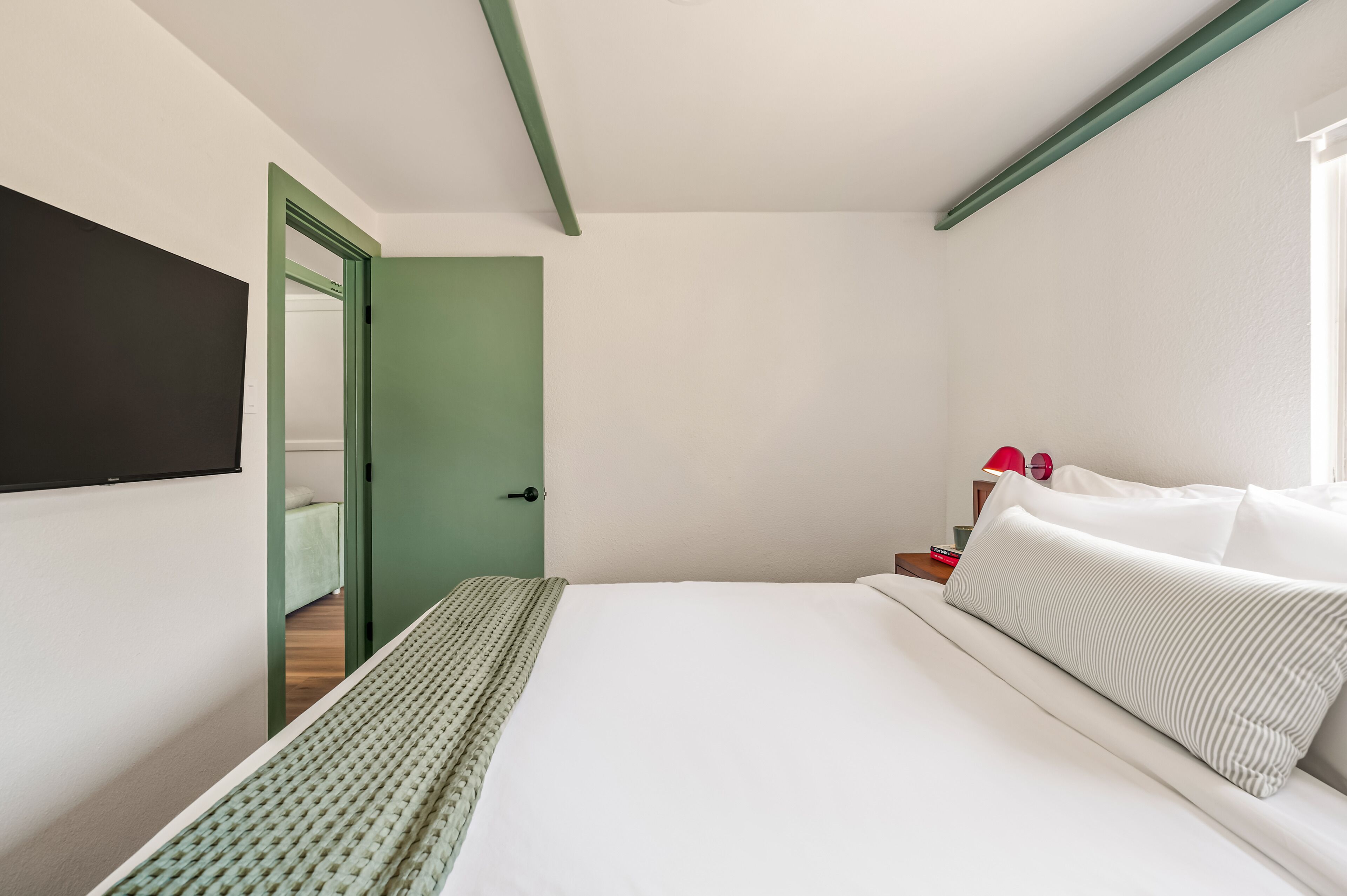 A modern hotel room with a bed, a green door, and a red lamp on a nightstand.