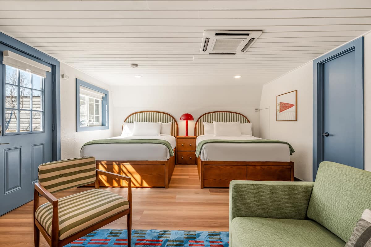 A bedroom with two beds, a chair, a sofa, and a window with blinds.