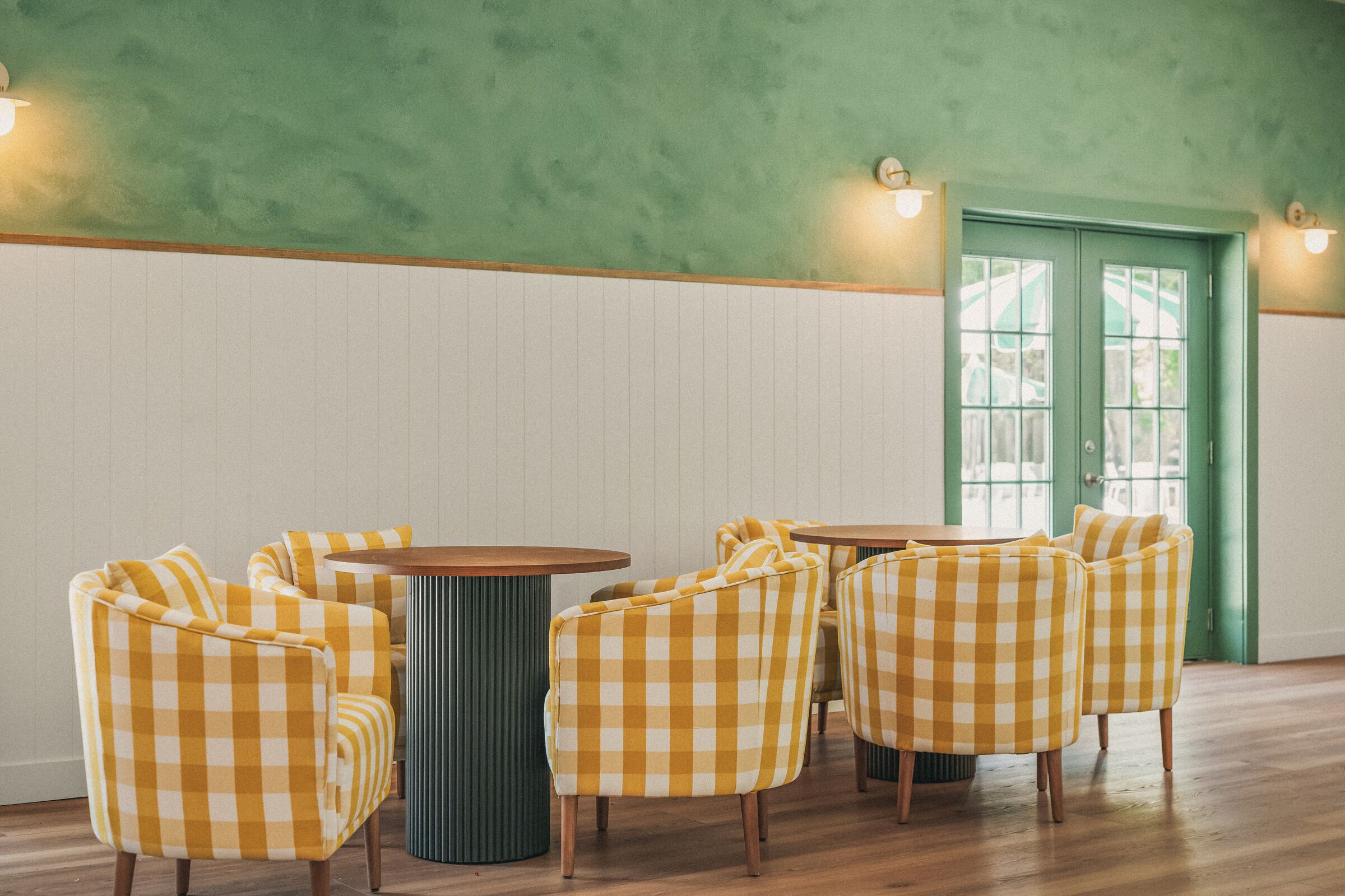 A room with green walls, white paneling, and a green door. There are yellow checkered chairs around a round table.