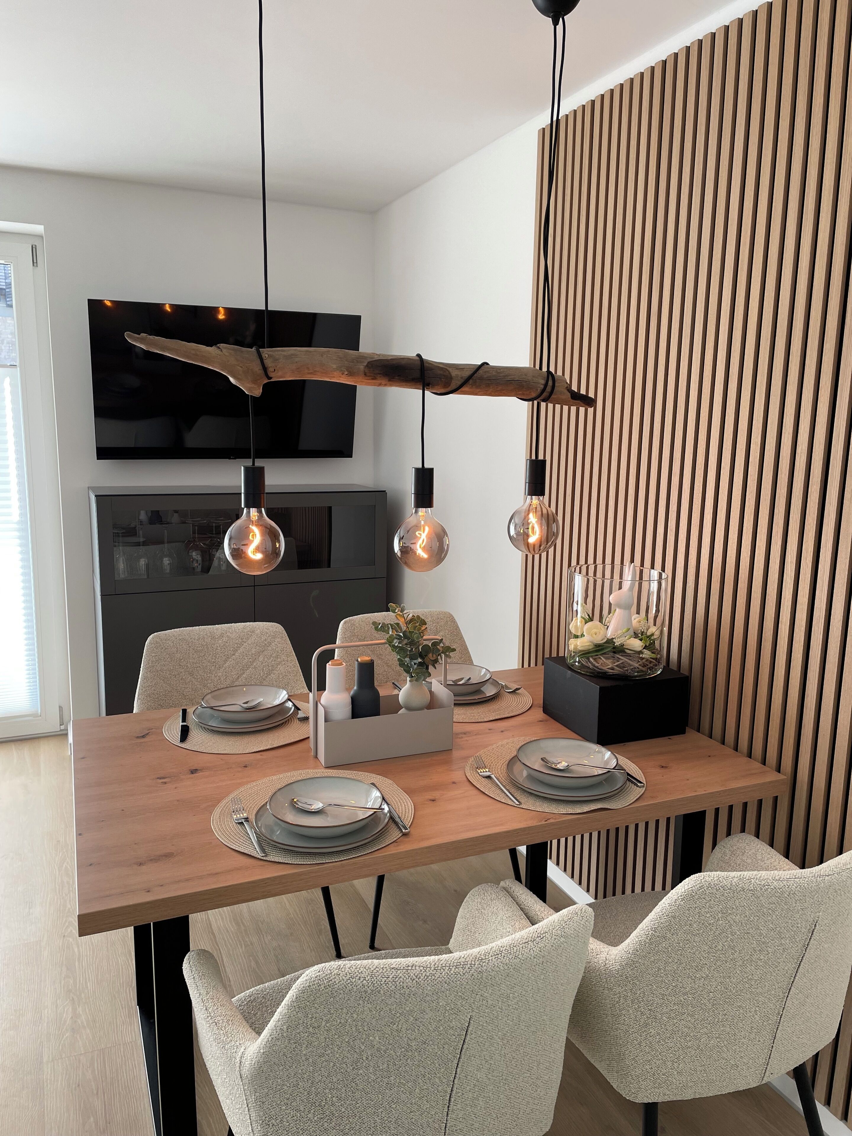 A modern dining area with a wooden table set for four, featuring a central vase with flowers, a black TV mounted above, and a wooden slat wall to the right.