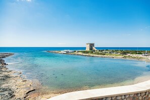 A coastal view with clear blue waters, a rocky shoreline, and a tower-like structure in the distance. - Hotel Alba (Torre Lapillo)