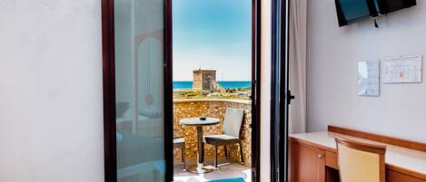 Quarto de hotel com cama, secretária, televisão e varanda com vista para o mar e uma torre.