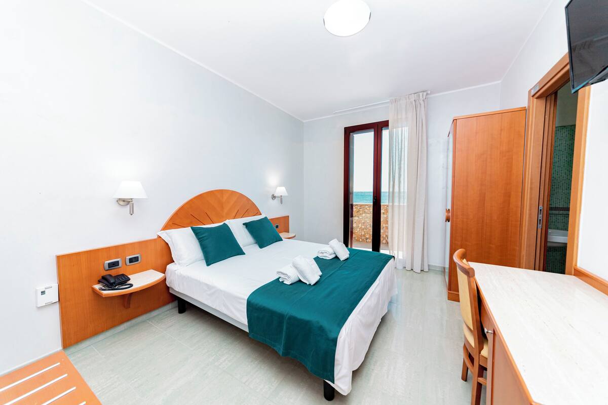 A hotel room with a bed, bedside tables, a desk, and a view of the sea.