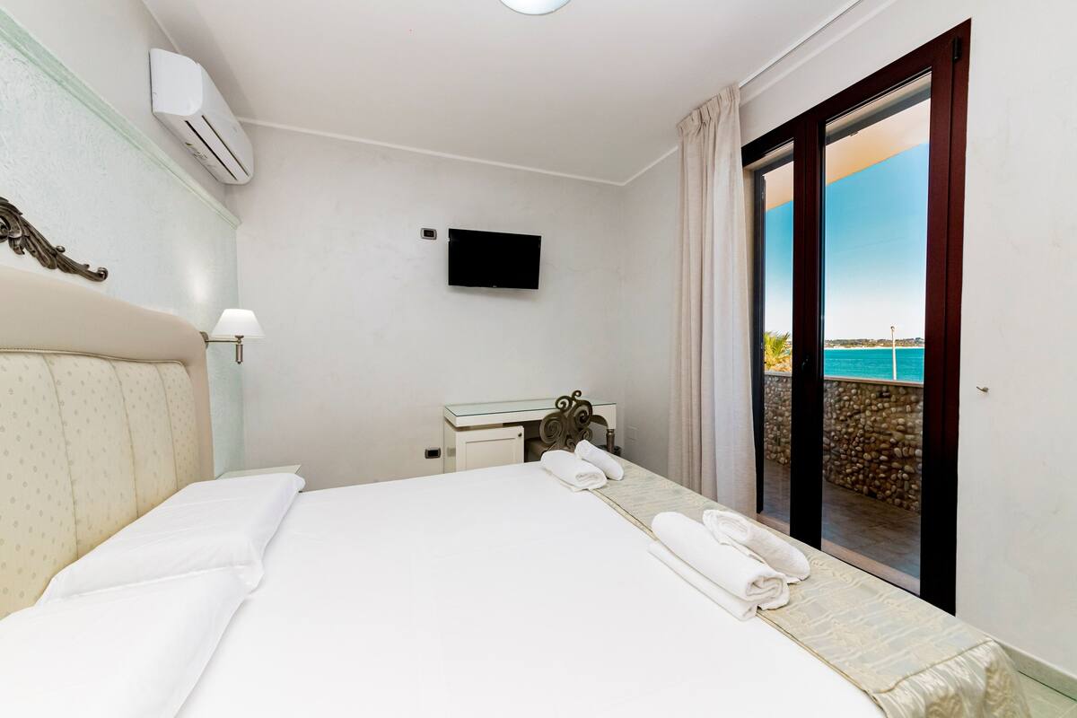 A hotel room with a bed, a TV, a desk, and a view of the sea.