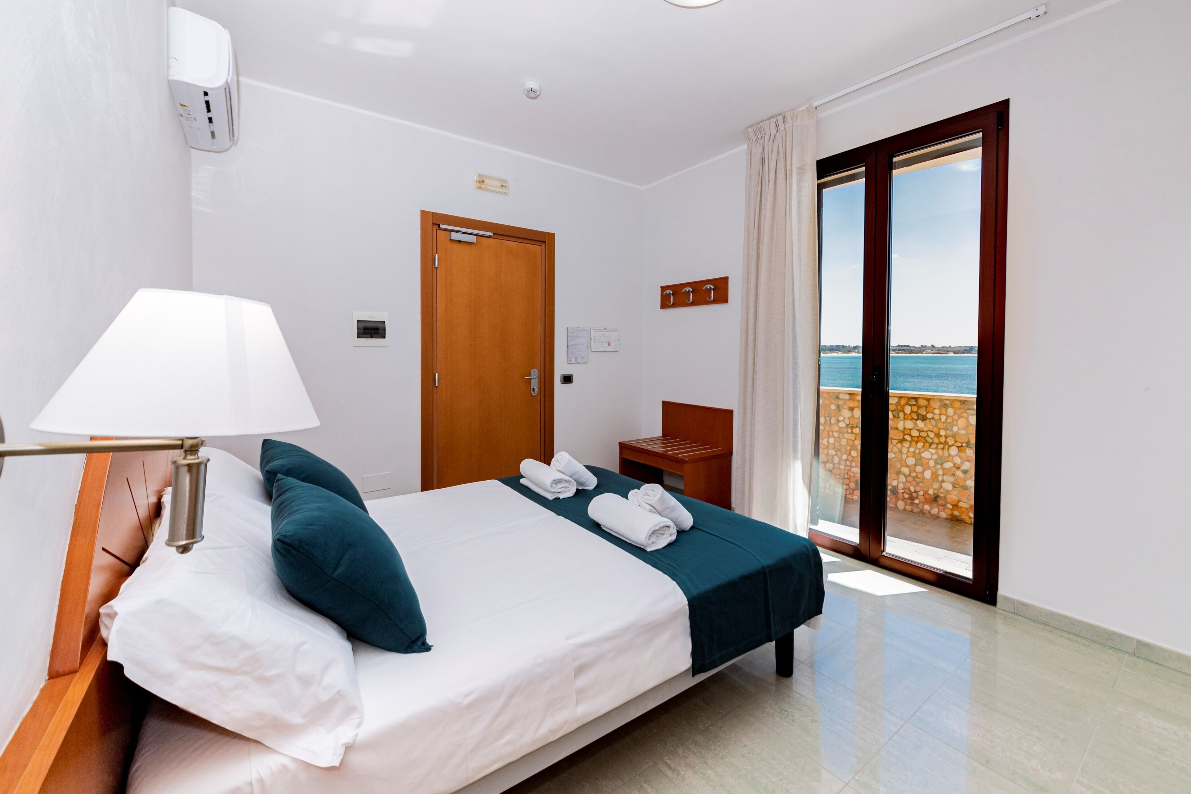 A hotel room with a bed, a nightstand, a lamp, a door, and a view of the sea through a sliding glass door.