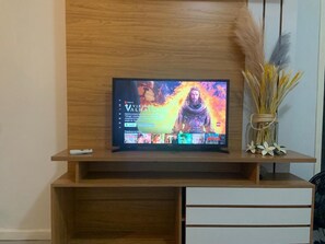 A television on a wooden TV stand with a remote control, a vase with dried plants, and a wall with a wooden panel. - Centro da Cidade dois quartos com ar (Cabo Frio)
