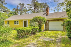 A yellow single-story house with a garage, surrounded by greenery and trees. - Bike to Cape Cod Sights: Home w/ Backyard Oasis (Wellfleet)