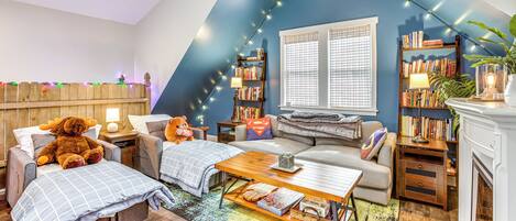 A cozy room with a loft bed, a sofa, a coffee table, and a fireplace. There are bookshelves and a window with blinds.