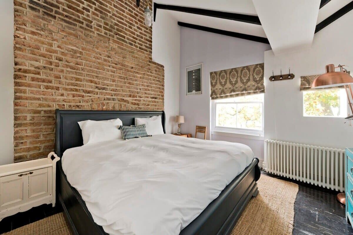 Un dormitorio con pared de ladrillo, cama con ropa de cama blanca, radiador y ventana con cortinas estampadas.