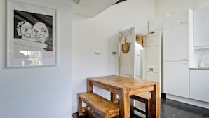 A modern kitchen with white cabinetry, a wooden table with benches, and a framed artwork on the wall. - 1 Bed Bohemian Loft Style Apt (London)