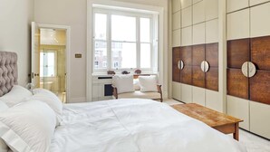 A bedroom with a large bed, a wooden bench, a window with a view, and a wardrobe with a unique design. - Grand 2 Bedroom Flat, Kensington (London)
