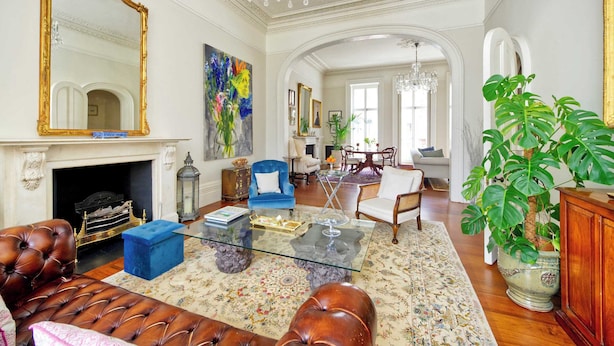 A spacious living room with a fireplace, a large mirror, and a chandelier. There is a blue armchair, a leather sofa, and a dining area in the background. - Grand 2 Bedroom Flat, Kensington (London)