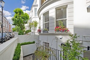 Une maison de ville blanche à la façade incurvée, un petit jardin agrémenté de plantes en pot et une vue sur la rue où sont garées des voitures.