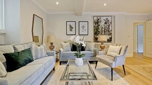 En moderne stue med en lyseblå sofa, en grå lænestol og et sofabord i glas med en hvid vase. To indrammede botaniske tryk hænger på væggen, og over sofaen er der et stort abstrakt maleri.