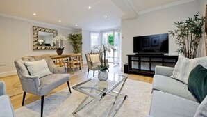 En moderne stue med sofa, lænestol og sofabord, et spiseområde med stole, et fjernsyn og et stort spejl.