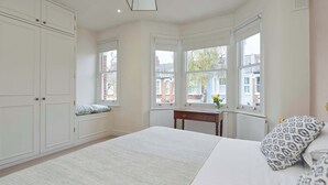 A bedroom with a large bed, a nightstand, a dresser, and a window with a view of the outside. - Central London 4BR Queens Park Family Home With Garden (London)