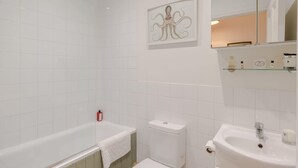 Un baño con bañera, inodoro y lavabo. Hay una toalla en la bañera, un dispensador de papel higiénico y un espejo sobre el lavabo.