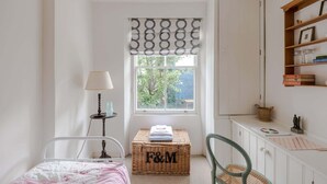 Un dormitorio con cama, una mesita de noche con lámpara, ventana con cortina estampada, una pequeña cesta de mimbre y un escritorio con estantes.