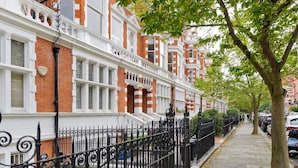 A row of terraced houses with white and red brick facades, black wrought iron fences, and a tree-lined street. - Spacious 3-bedroom Apartment in Earls Court (London)