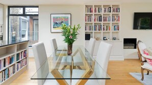A modern living room with a glass dining table, white chairs, a bookshelf, and a fireplace. - Spacious 3-bedroom Apartment in Earls Court (London)