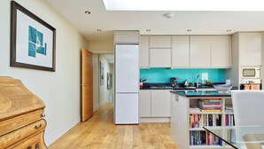 A modern kitchen with wooden flooring, a white refrigerator, and a teal backsplash. There is a bookshelf with books and a dining area with a glass table. - Spacious 3-bedroom Apartment in Earls Court (London)