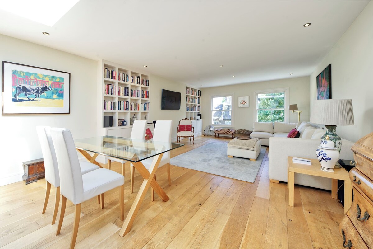 A spacious living room with a glass dining table, white sofas, and a bookshelf filled with books.