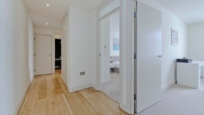 Un couloir au plancher de bois mène à une chambre à coucher qui contient un lit et une petite table blanche.