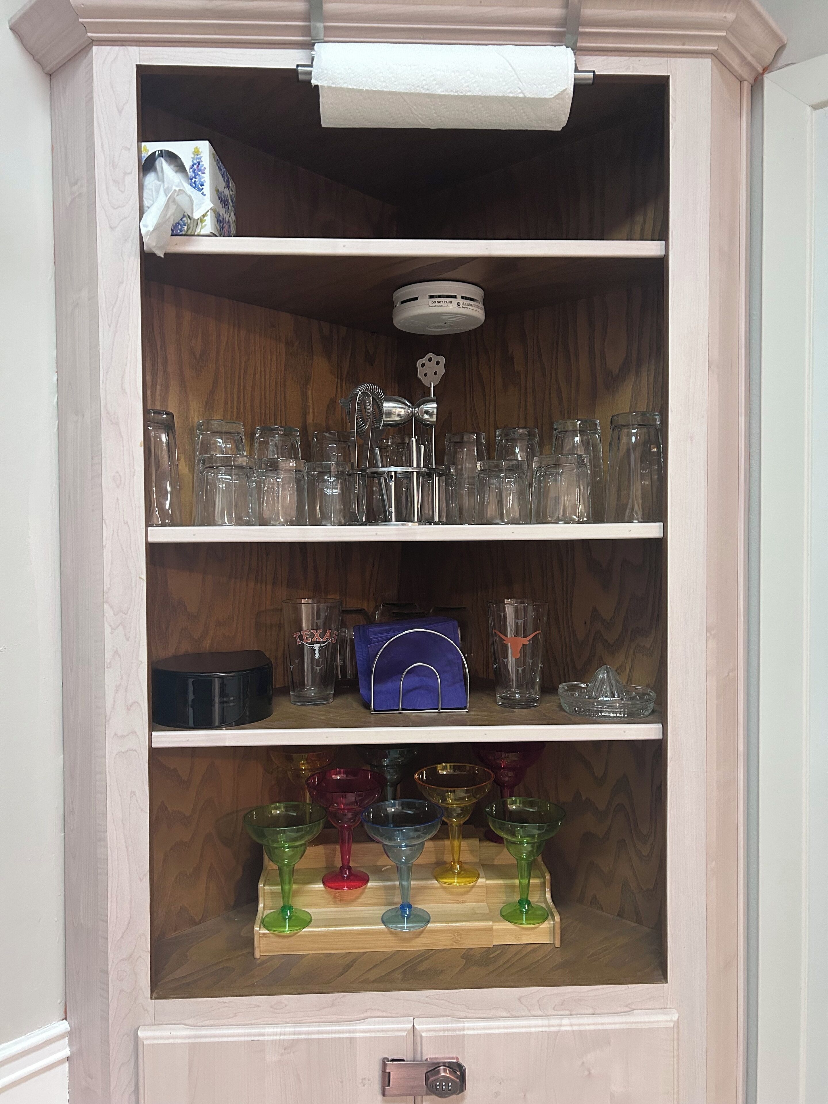 A wooden cabinet with shelves holding glassware, a blue bag, and a Texas-themed glass.