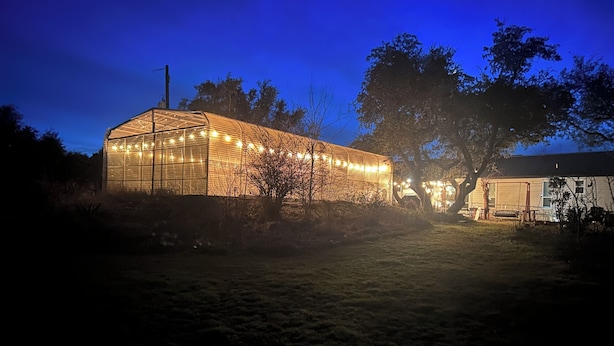 A large, illuminated greenhouse structure with a metal frame and glass panels, surrounded by trees and a grassy area. - Spacious country oasis:Swim,BBQ, Attend Weddings!Close to venues in Hill Country (Austin)