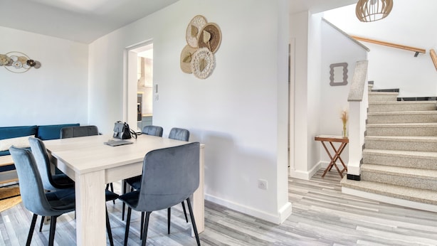 Un comedor moderno con una mesa de madera y sillas grises tapizadas, una escalera con barandilla de madera y un suelo de tono claro.