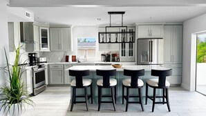 Cocina moderna con muebles blancos, una isla central con taburetes altos, electrodomésticos de acero inoxidable y un amplio ventanal que proporciona luz natural.