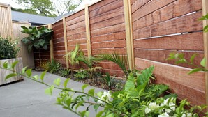 A wooden fence with a slatted design, surrounded by various plants and a concrete pathway.