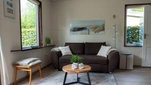A living room with a brown sofa, a wooden coffee table with two white vases, a small wooden stool, and a large window with a view of greenery. - Jamies Cottage Warmenhuizen (Warmenhuizen)