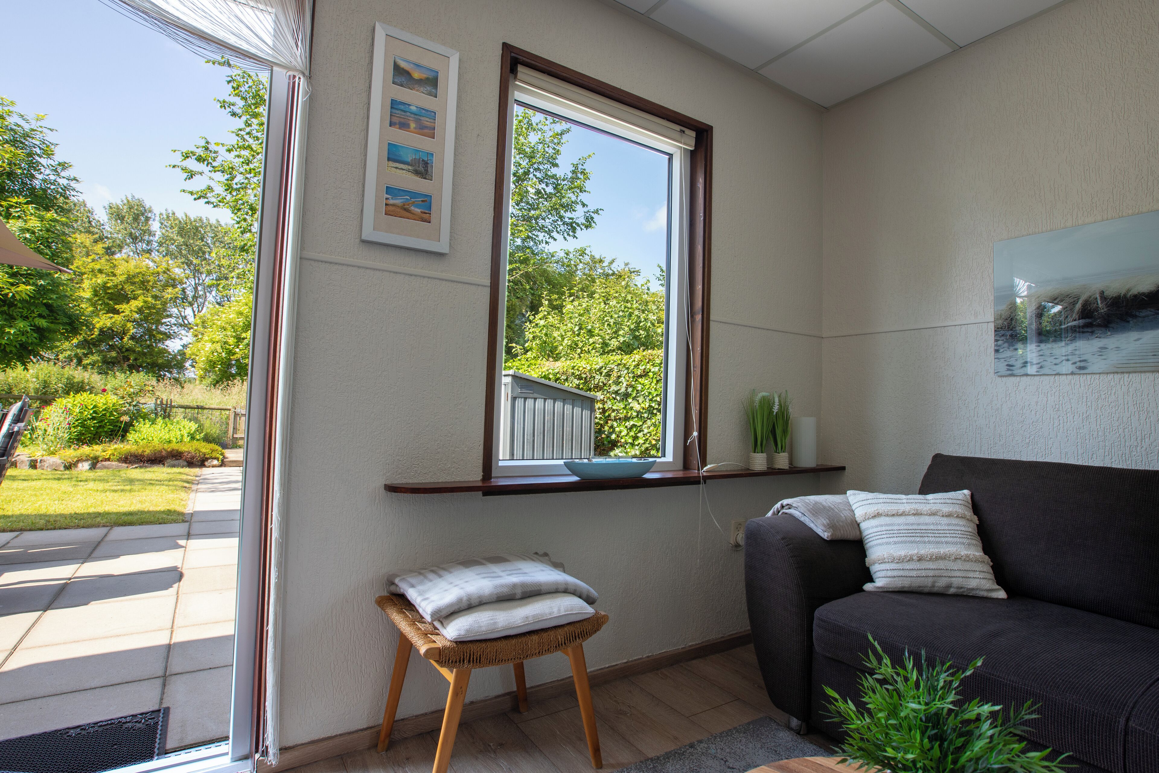 Un salon avec un canapé foncé, un petit tabouret, une fenêtre donnant sur un espace verdoyant et un tableau encadré accroché au mur.