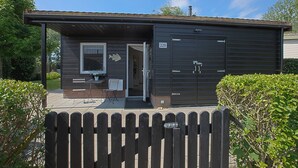 A small, single-story building with a dark exterior, a visible number "228" on the door, and a small porch area with a chair and table. - Jamies Cottage Warmenhuizen (Warmenhuizen)