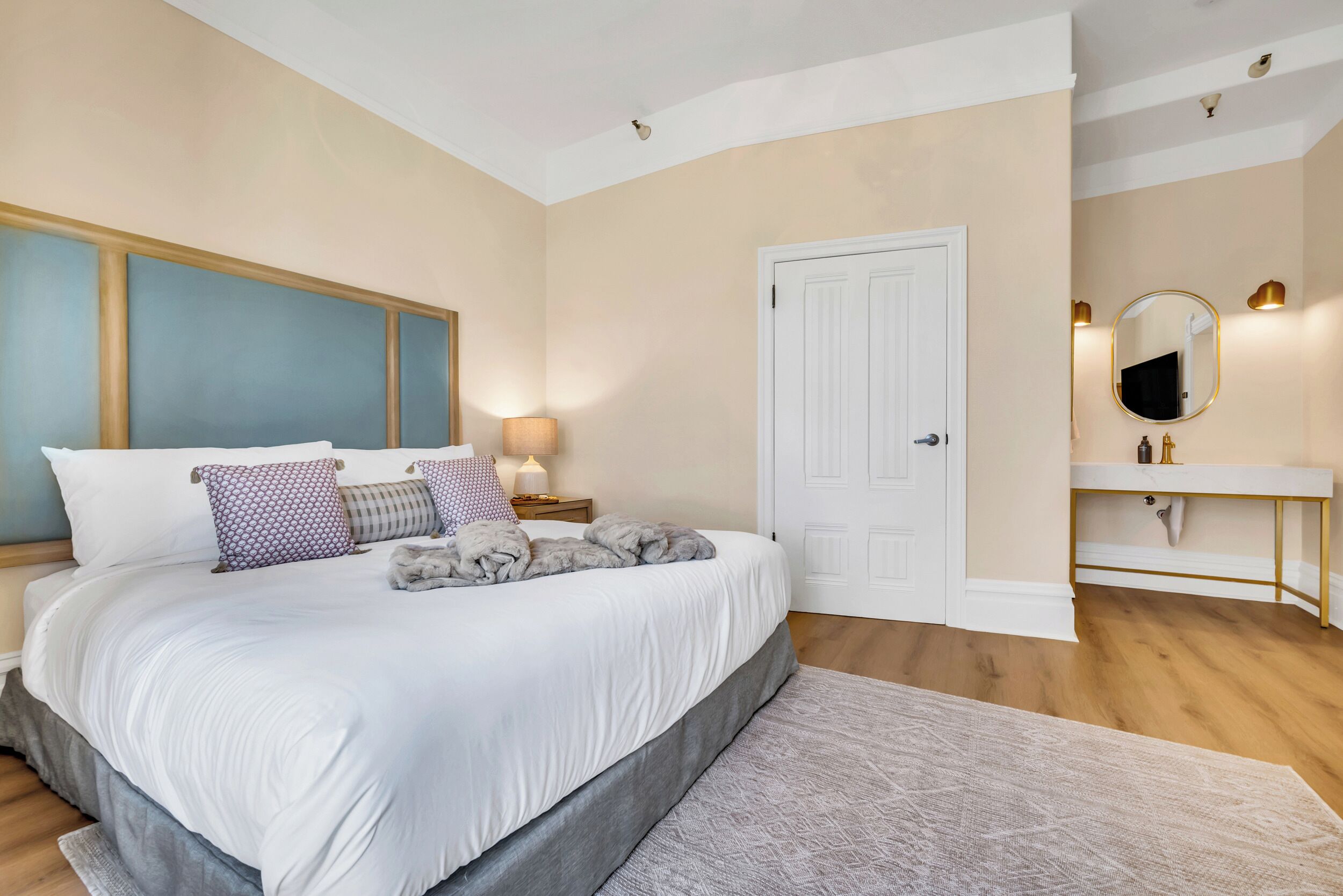 Un dormitorio con una cama grande, una mesita de noche con lámpara, una puerta blanca y un baño con espejo y lavamanos.