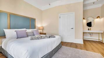 Un dormitorio con una cama grande, una mesita de noche con lámpara, una puerta blanca y un baño con espejo y lavamanos.