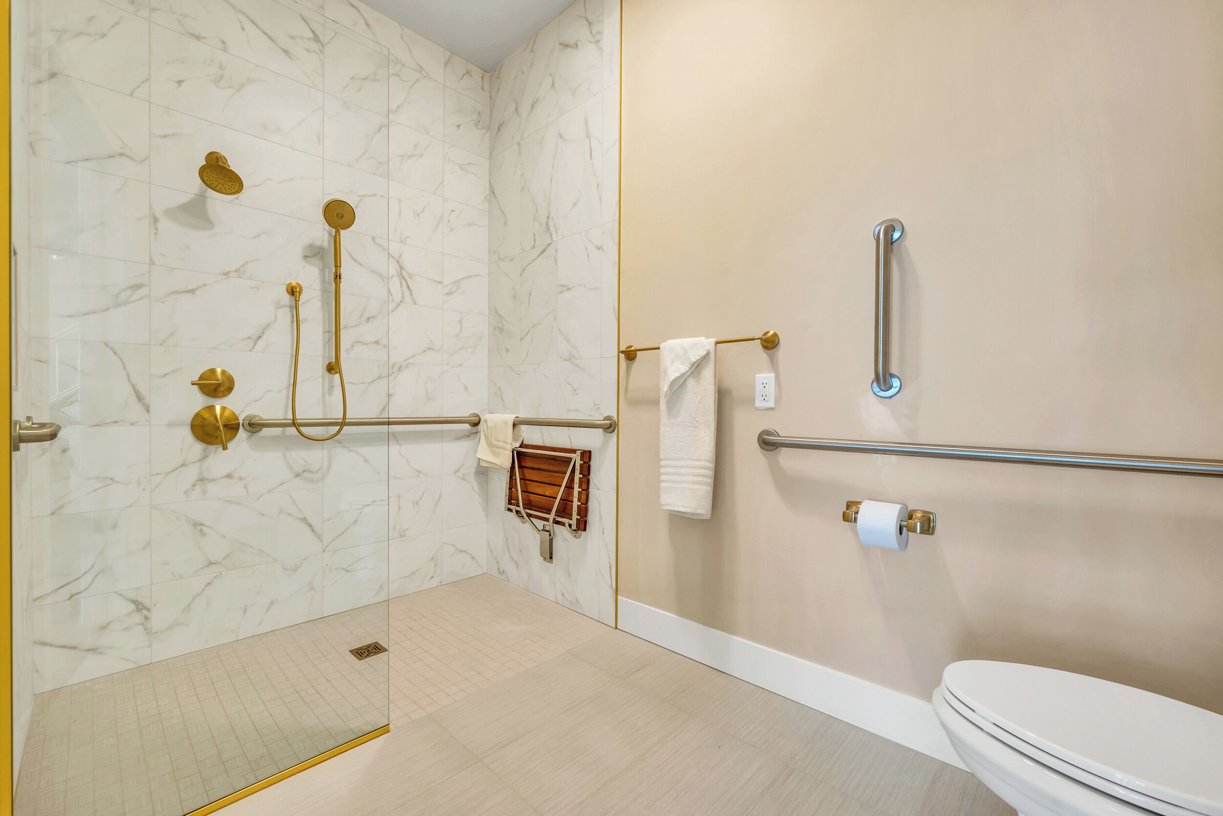 A bathroom with a marble wall, a toilet, a shower with gold fixtures, and a grab bar.