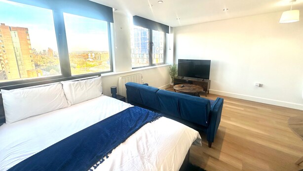 A modern hotel room with a large bed, a seating area with a TV, and large windows offering a city view. - Up to 40 Percent Off Old Trafford Modern Apartment (Manchester)