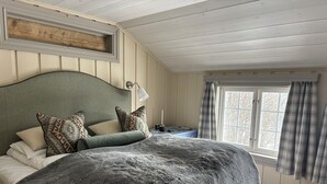 A bedroom with a large bed, a headboard, a bedside table with a lamp, a window with curtains, and a wooden ceiling.