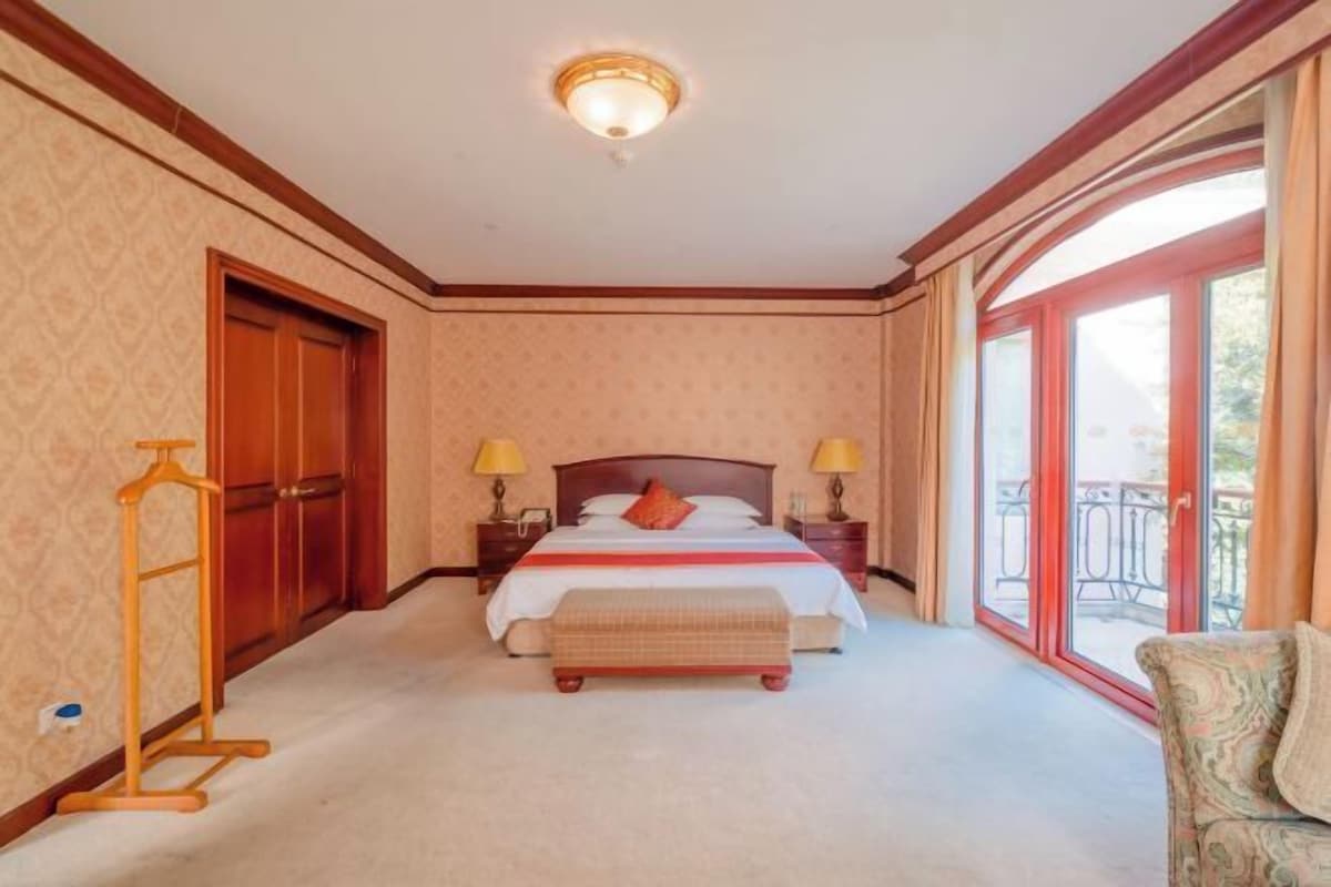 A hotel room with a bed, bedside lamps, a wooden wardrobe, a bench, and a balcony with a railing.