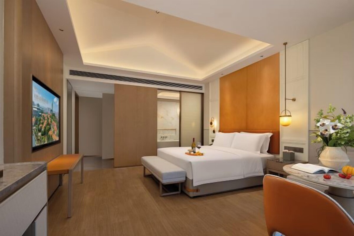 A hotel room with a bed, a desk, a chair, a television, and a wall-mounted lamp.