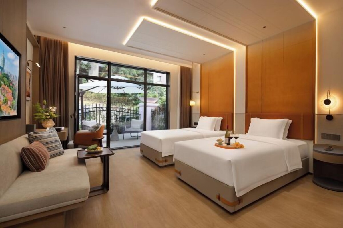 A hotel room with a sofa, two beds, a small table, and a balcony with outdoor furniture.