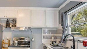 A compact kitchen with white cabinets, a stainless steel stove, and a window with a view of trees. - Cozy 3-Bedroom Home – Sleeps 6 (Tulsa)