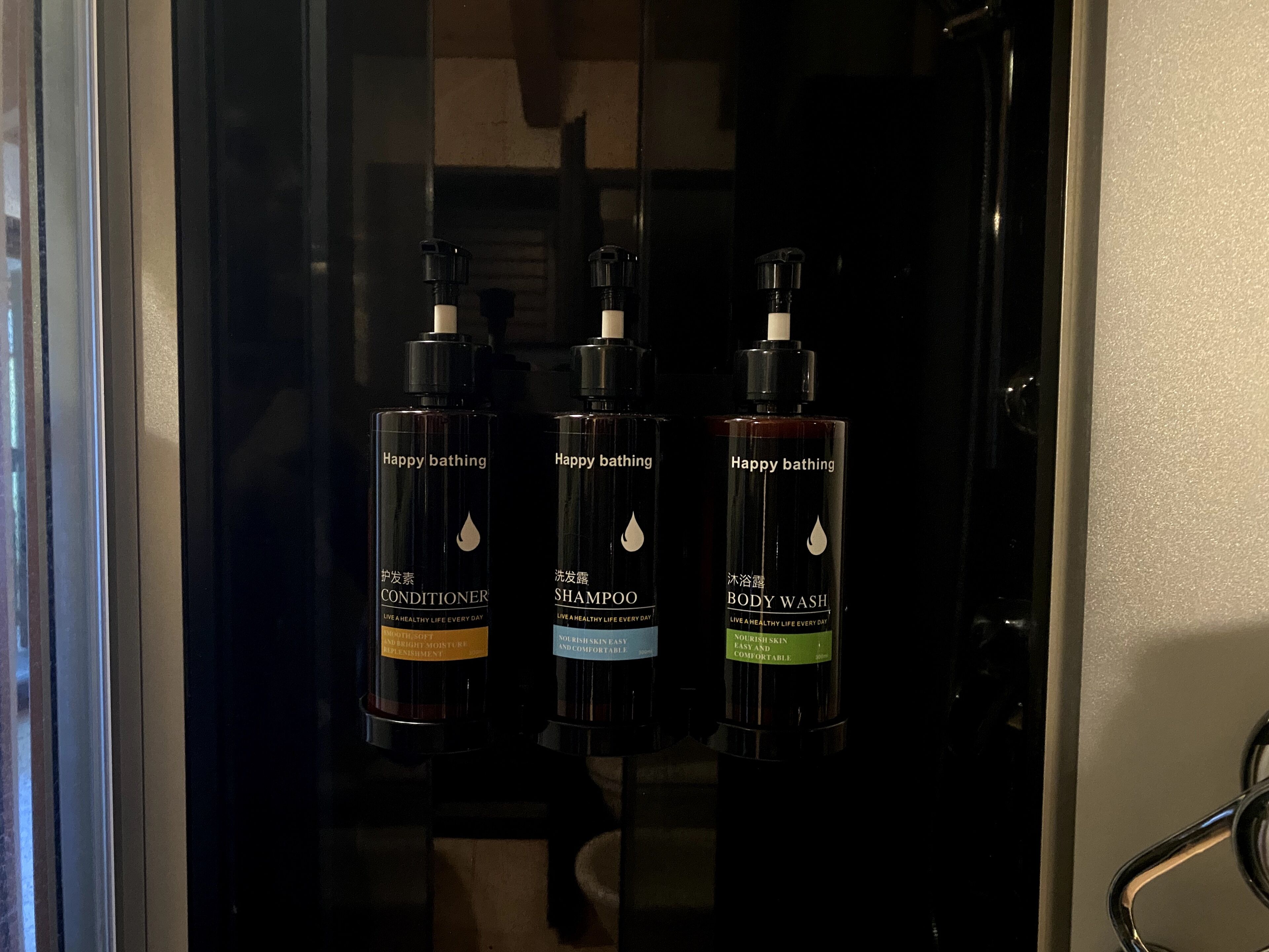 Trois flacons de produits de bain de la marque Happy : après-shampoing, shampoing et gel douche, présentés dans une pièce sombre.