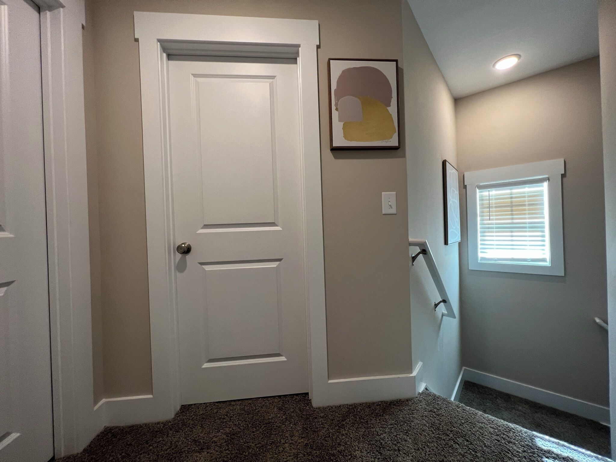 A hallway with a white door, a framed abstract painting, a window with blinds, and a carpeted floor.