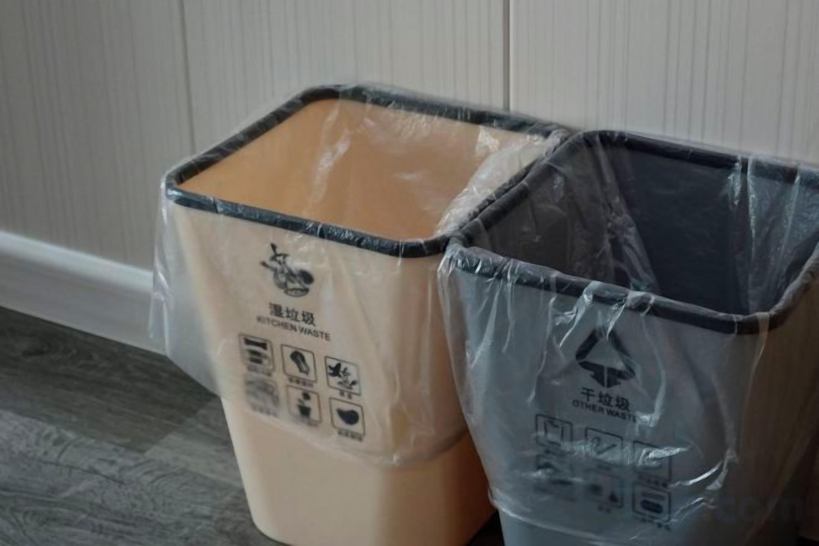 Two trash bins, one beige and one black, with recycling symbols and text on them, placed against a wall.