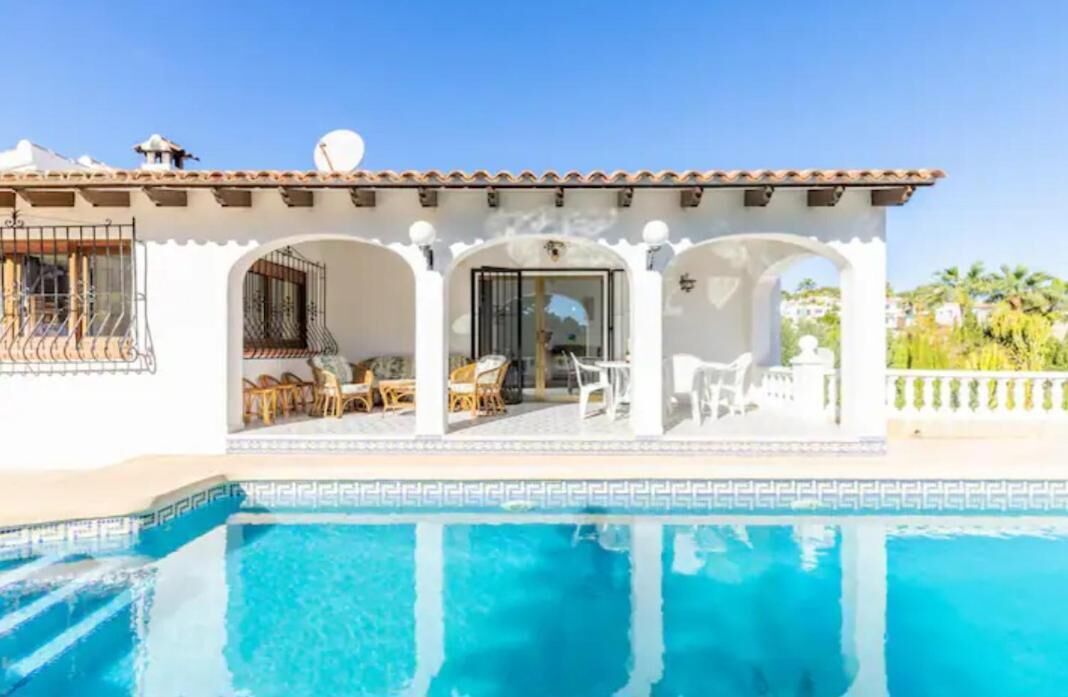 A white building with a pool, featuring a patio with chairs and tables, and a clear blue sky.