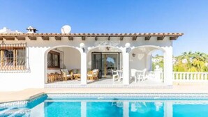A white building with a pool, featuring a patio with chairs and tables, and a clear blue sky.