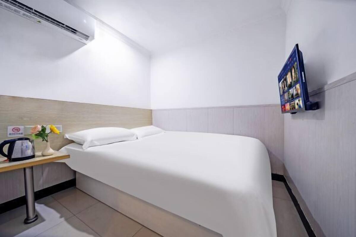 A hotel room with a bed, a small table with a kettle, a vase with flowers, and a mounted TV.